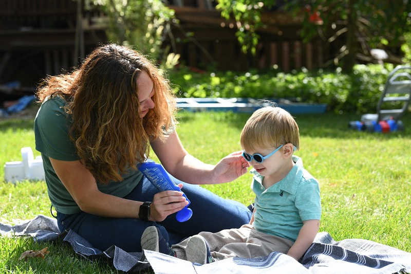 Mother putting sunscreen on her young son while sitting out in the grass on a warm sunny day.