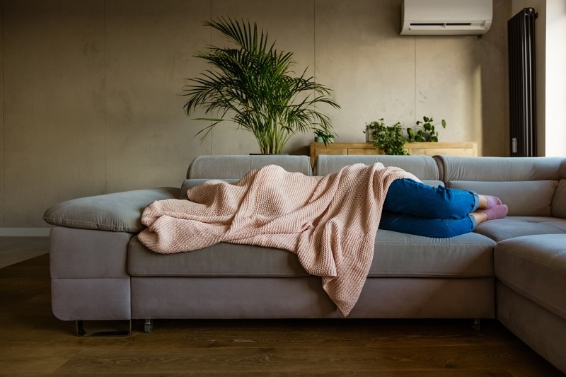  Young woman lying down on sofa in living room covered by blanket.
