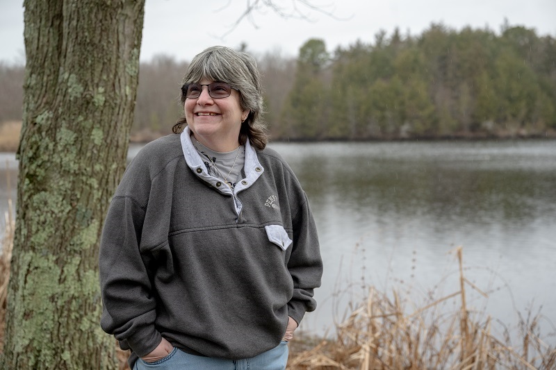 Woman, Elly Levine, standing in front of a lake smiling.