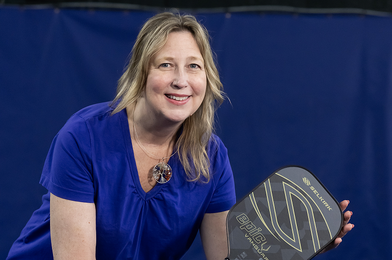 Diane Blaszka holding a pickleball racket, ready to play and smiling. 