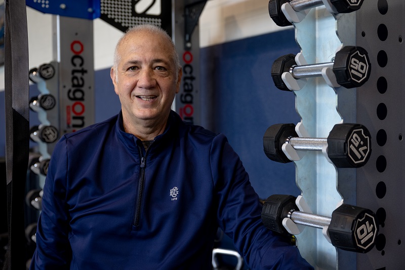 Anthony Lucia smiling at the gym, standing in front of some weights.