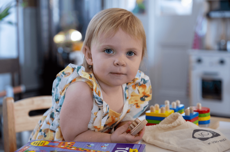 Toddler Marigold Mackey leans over a table covered in toys while looking at the camera.